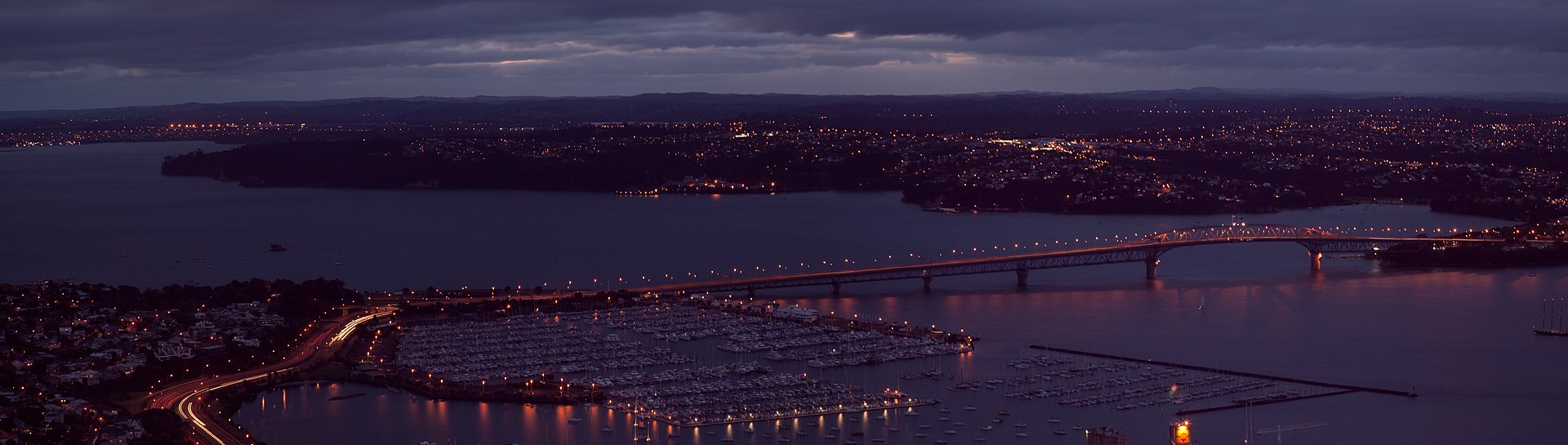 Auckland Harbour Bridge at dusk, city lights coming on across the Waitematā